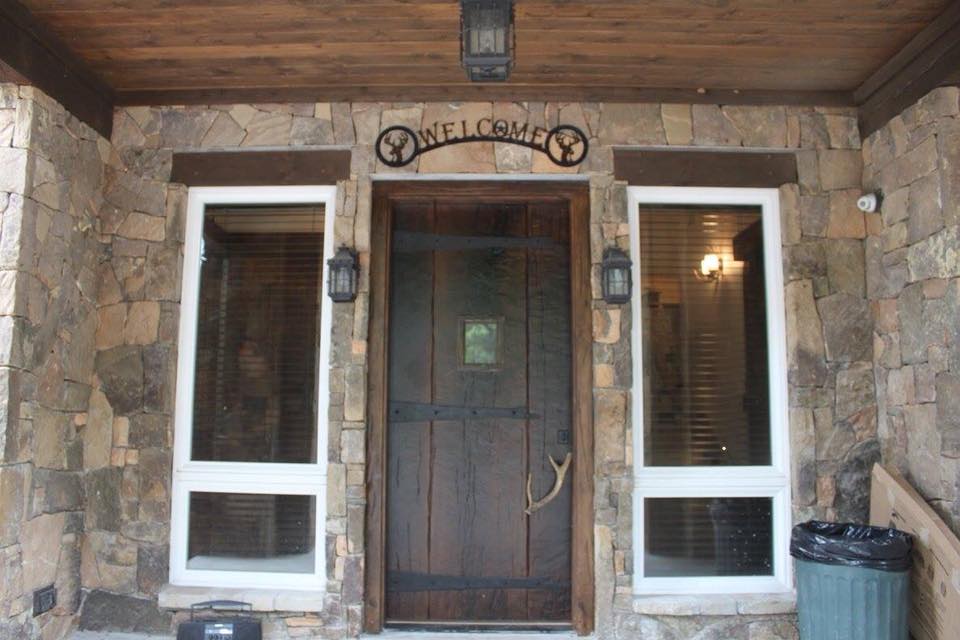 Stone entry porch with cedar beam ceiling, rustic wood door with iron Welcome sign and antler handle — Tincher's Custom Homes outdoor craftsmanship